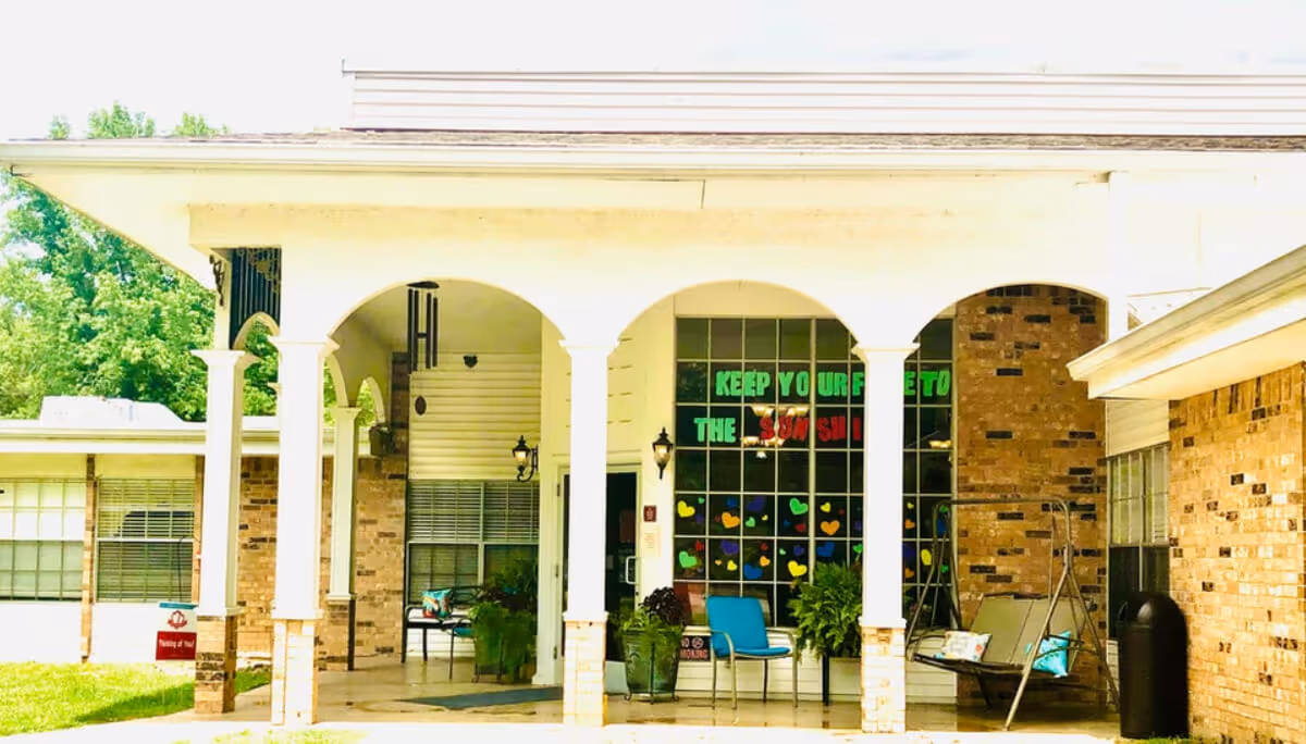 Entrance area of a senior living facility with a covered porch supported by white columns. There are chairs and potted plants on the porch, and a large window with colorful decorations and the message 'KEEP YOUR FACE TO THE SUNSHINE'. The building is made of light-colored brick and surrounded by greenery.