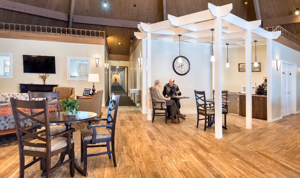 A bright and spacious common area in a retirement community featuring wooden floors, a round table with chairs, a seating area with a couch and armchairs, and two elderly men sitting and talking at a small table under a white pergola structure. A clock hangs on the wall behind them, and pendant lights illuminate the space. A hallway extends into the background.