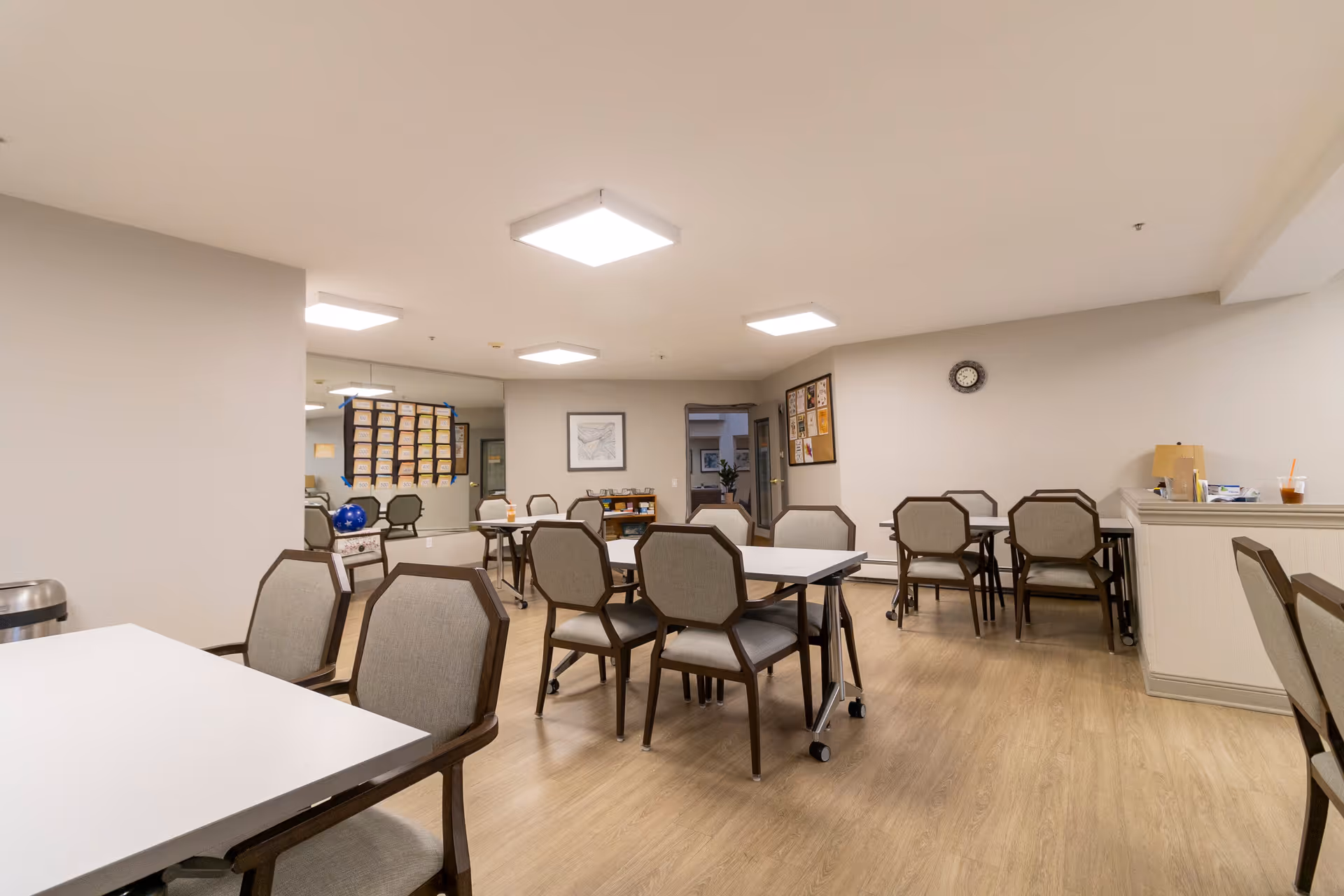 A well-lit common room with multiple tables and chairs arranged for group seating. The room has light-colored walls and wood flooring, with a clock on the wall and bulletin boards displaying various notices. There is a counter on the right side with some items on it, and a large mirror on the left wall reflecting part of the room.