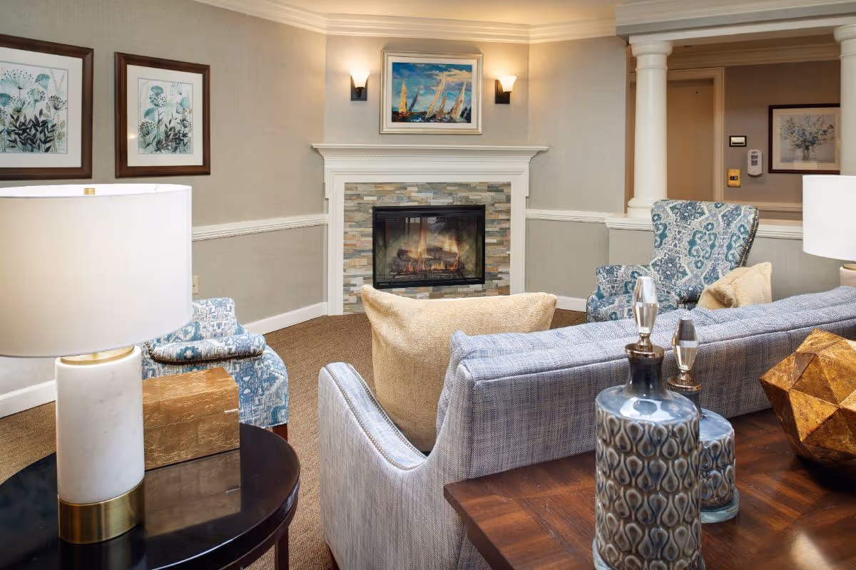 A cozy living room area with a lit fireplace surrounded by stone tiles and a white mantel. The room features a light blue sofa with beige cushions, two patterned armchairs, a side table with a white lamp, decorative vases on a wooden table, and framed artwork on the walls. The walls are beige with white trim, and there are two wall sconces above the fireplace providing warm lighting.