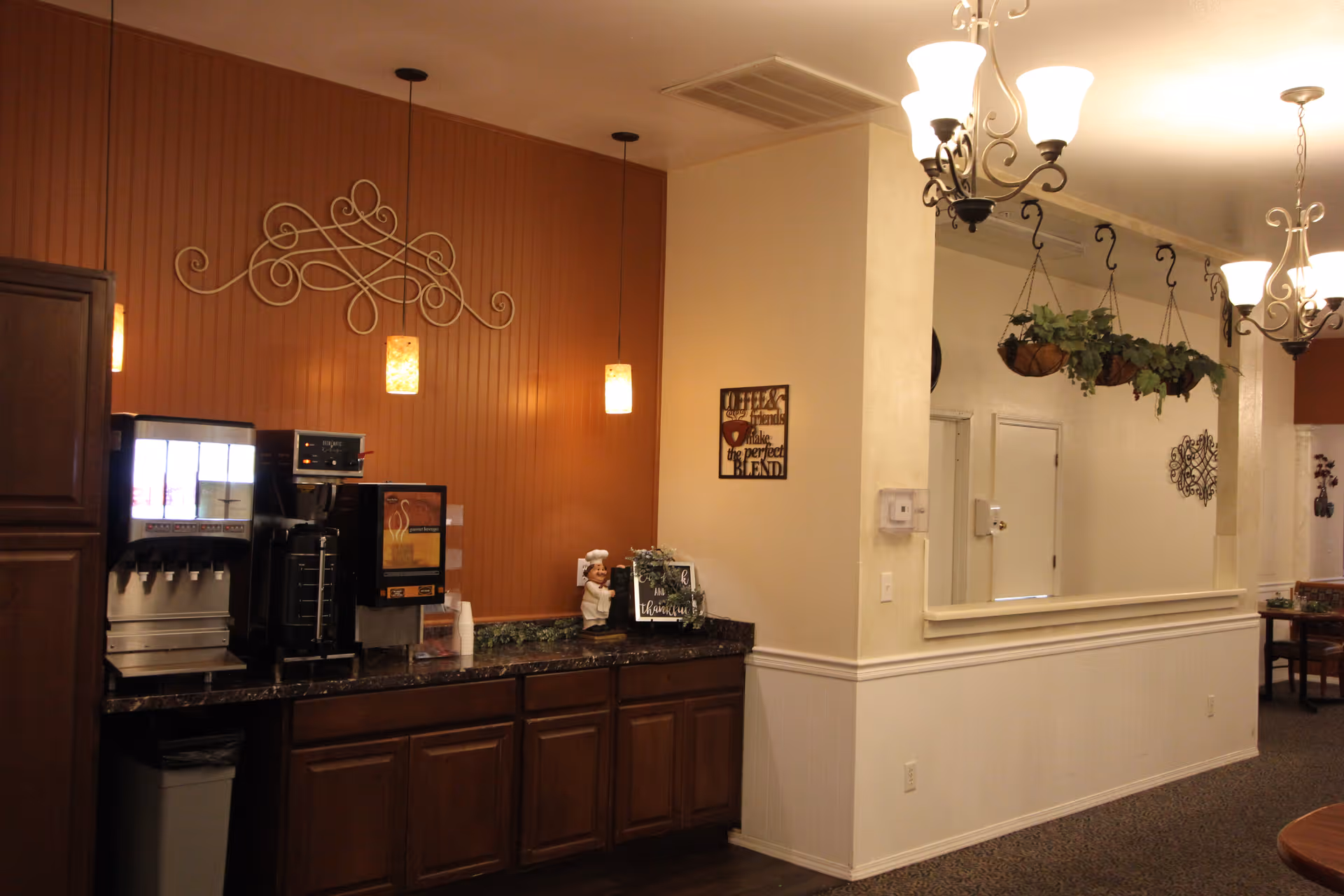 A coffee and beverage station in a retirement facility with a dark granite countertop, coffee machines, and a soda dispenser. The wall behind the station is painted a warm brown color with decorative wall art and hanging pendant lights. To the right, there is a large mirror with hanging plants above it and chandeliers on the ceiling.