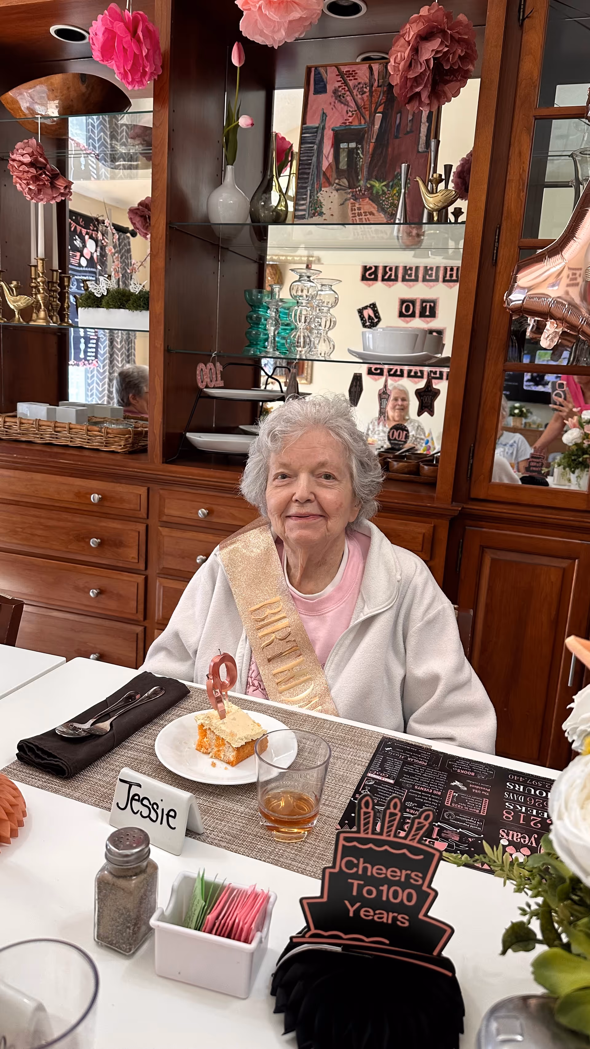 An elderly woman wearing a birthday sash sits at a dining table with a slice of cake and a drink in front of her. The table has a name card labeled 'Jessie' and a decorative sign that reads 'Cheers To 100 Years'. The background shows wooden cabinets with glass shelves and various decorative items.