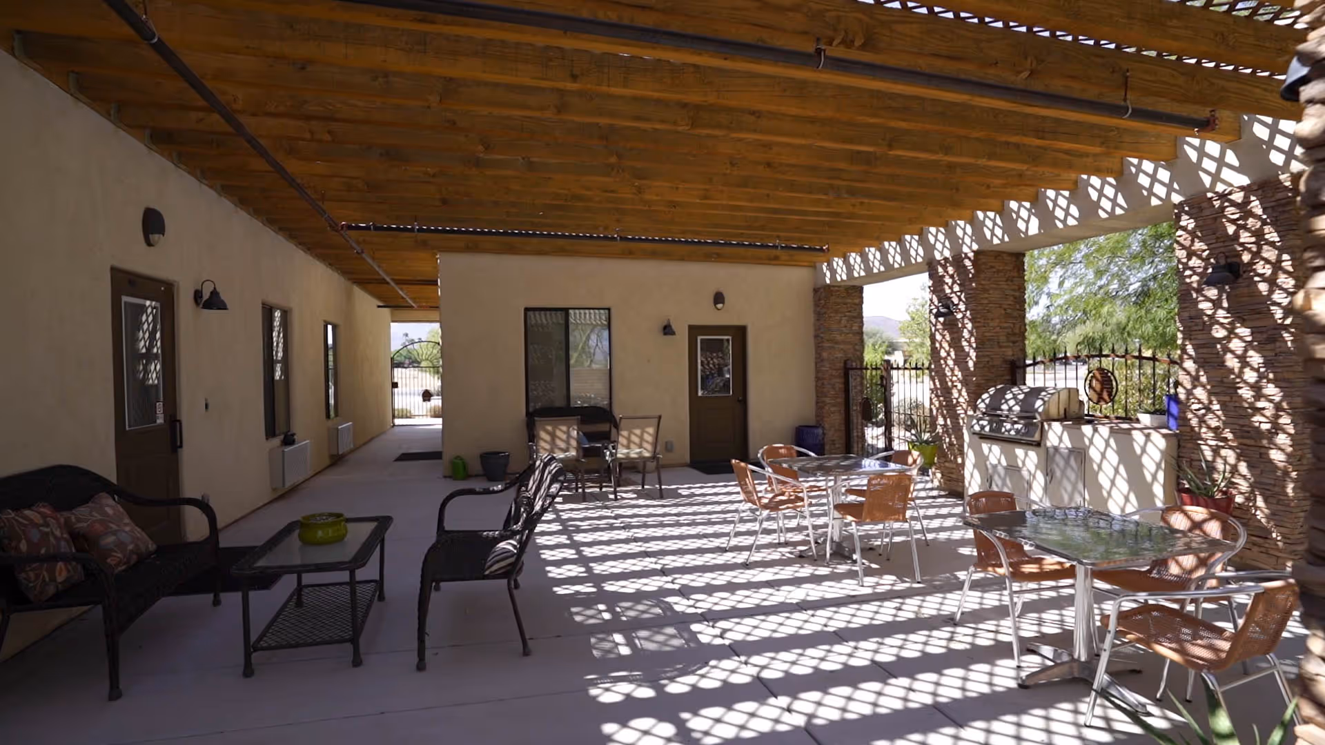 Covered outdoor patio area with wooden beams casting patterned shadows on the floor. The space includes several tables with chairs, a seating area with cushioned chairs and a coffee table, and a built-in barbecue grill. Stone pillars and a gated fence are visible, along with some greenery in the background.