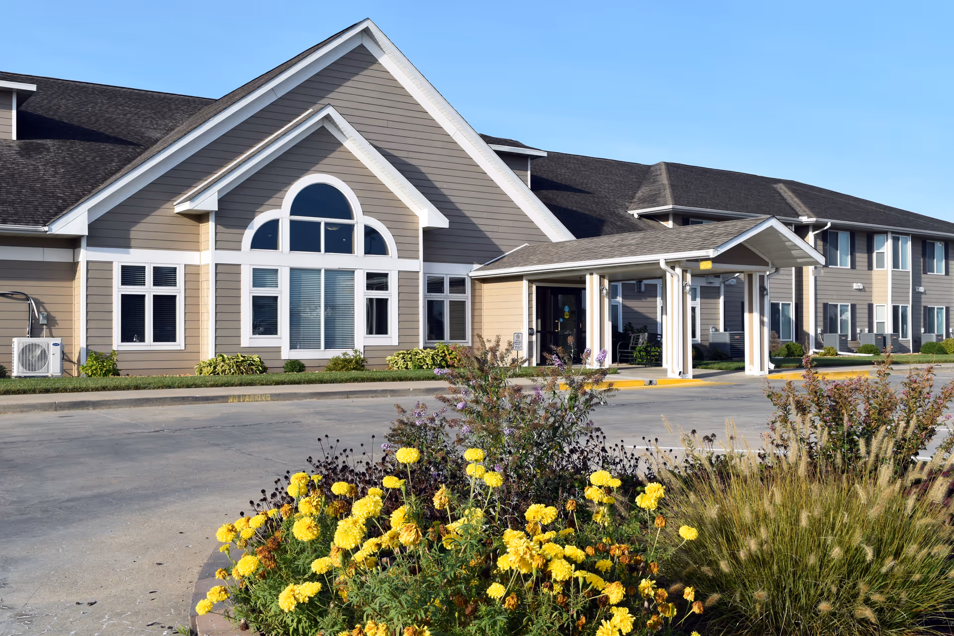 Exterior view of a two-story retirement community building with beige siding and white trim. The building features a covered entrance and large windows with decorative arches. In the foreground, there is a landscaped area with yellow flowers and ornamental grasses under a clear blue sky.