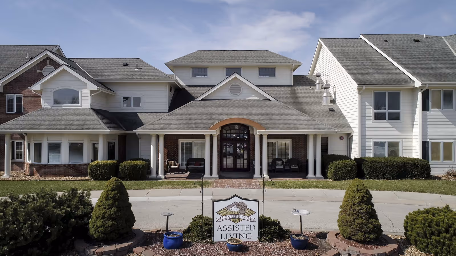 Front entrance of a two-story assisted living building with columns, landscaping, and a sign reading 'Assisted Living' in the foreground.
