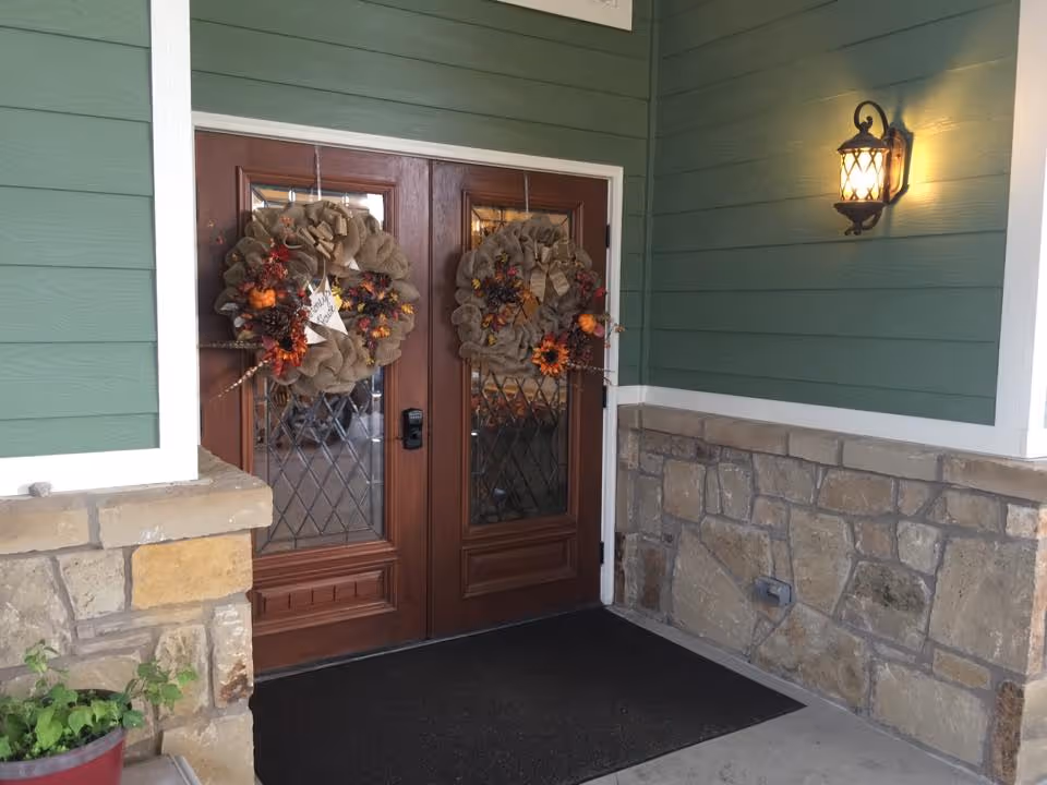 Double wooden front doors with glass panels decorated with autumn-themed wreaths, set in a green exterior wall with stone accents and a lit wall-mounted lantern on the right side.