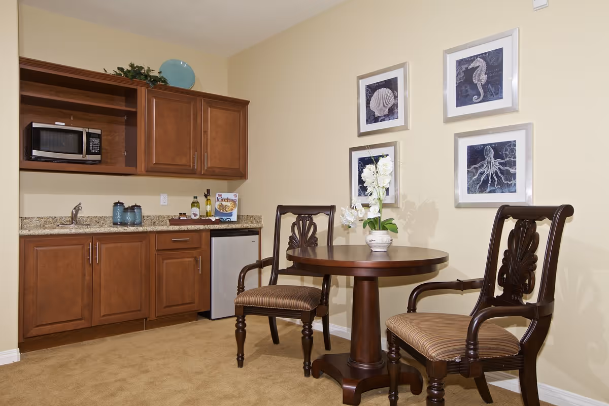 A small dining area with a round wooden table and two ornate wooden chairs with striped cushions. On the table is a white vase with white flowers. Behind the table is a wall with four framed pictures of sea creatures including a seahorse, shell, and octopus. To the left is a kitchenette with wooden cabinets, a granite countertop, a small sink, a microwave, and a mini refrigerator.
