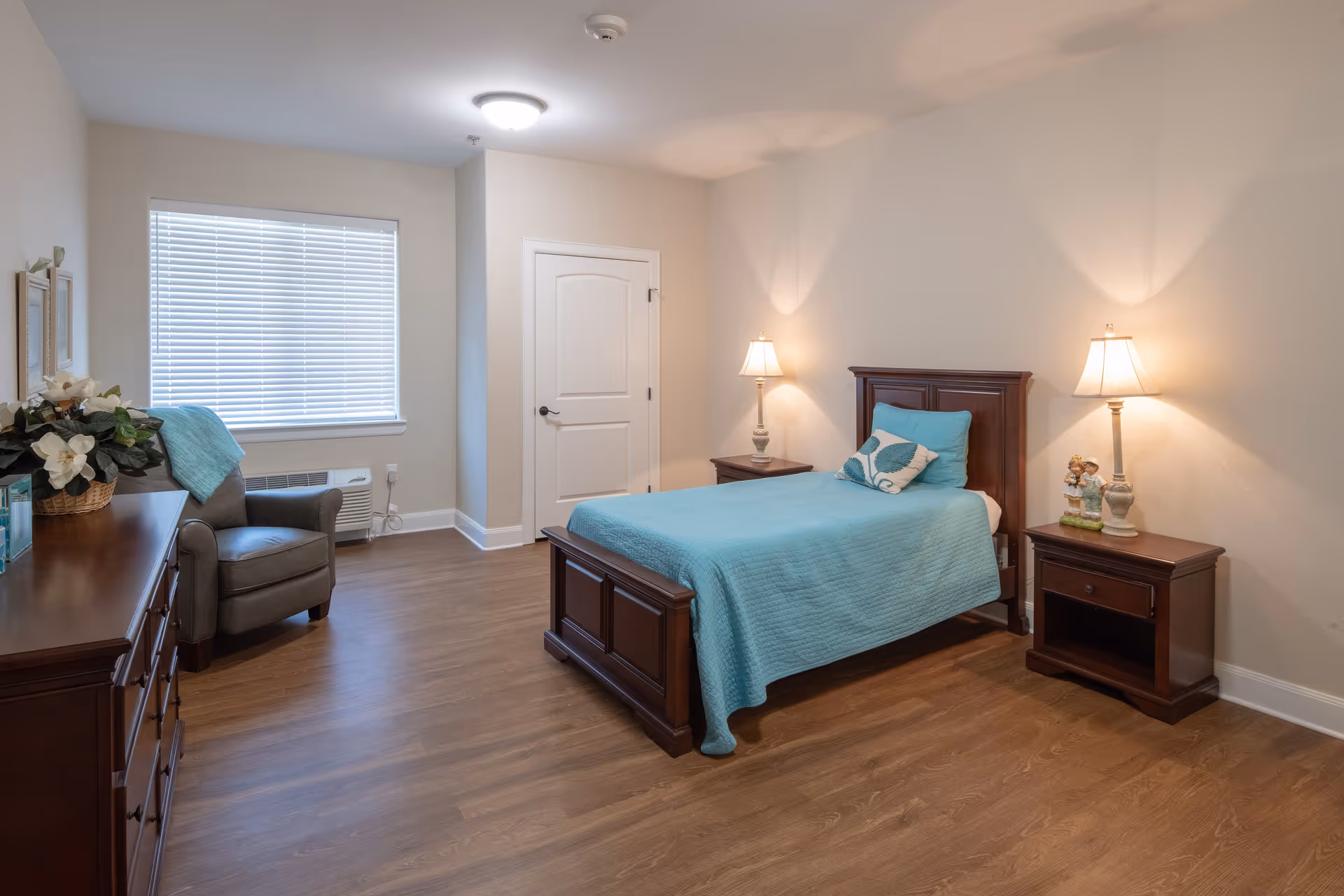 A senior living bedroom with a single wooden bed covered with a turquoise quilt and matching pillows. There are two wooden nightstands on either side of the bed, each with a lamp. A wooden dresser with a flower arrangement and a gray armchair are positioned near a window with closed blinds. The room has wood flooring and beige walls.