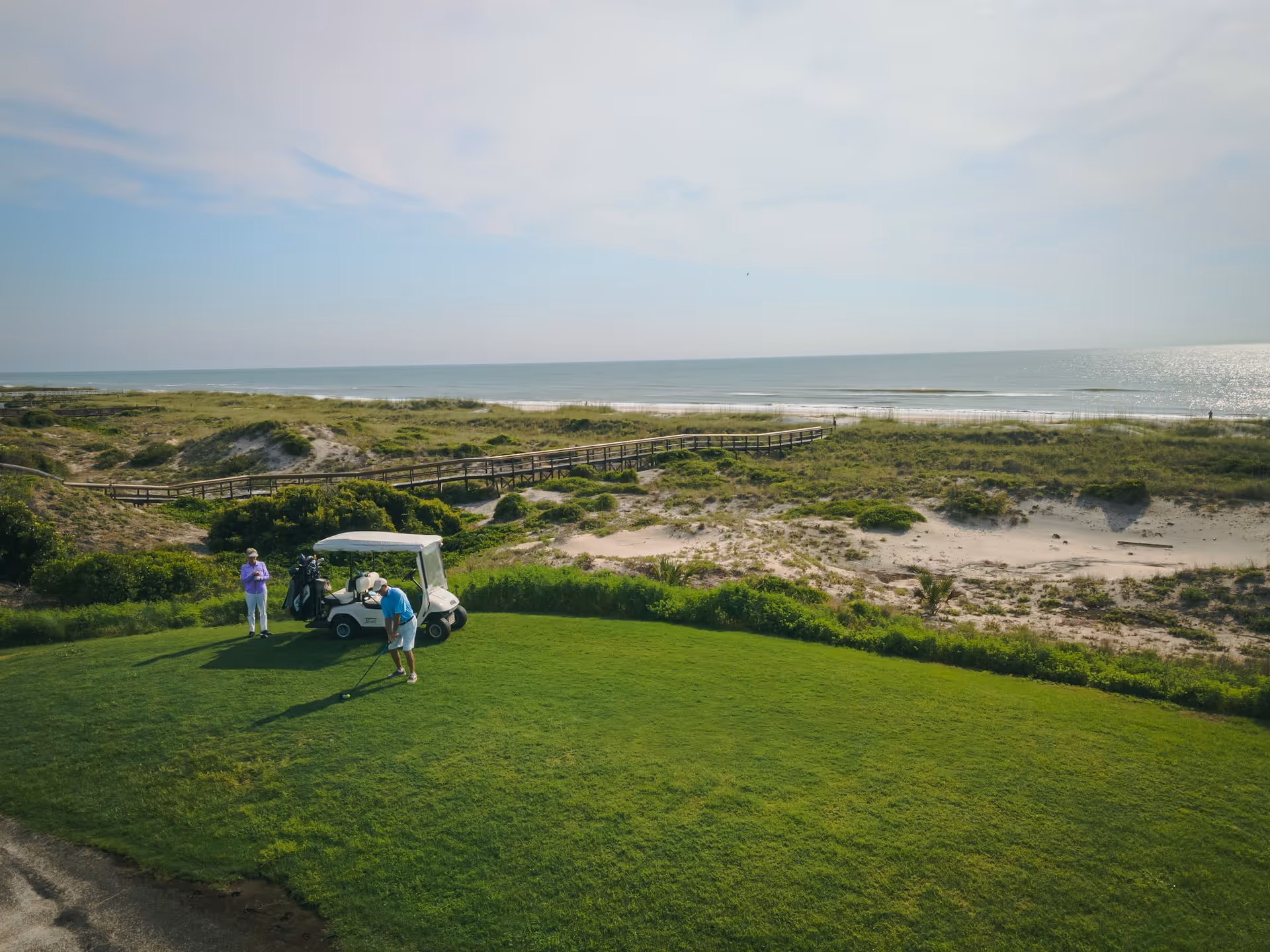 Two people playing golf on a green lawn near a golf cart with the ocean and sandy dunes in the background under a partly cloudy sky.