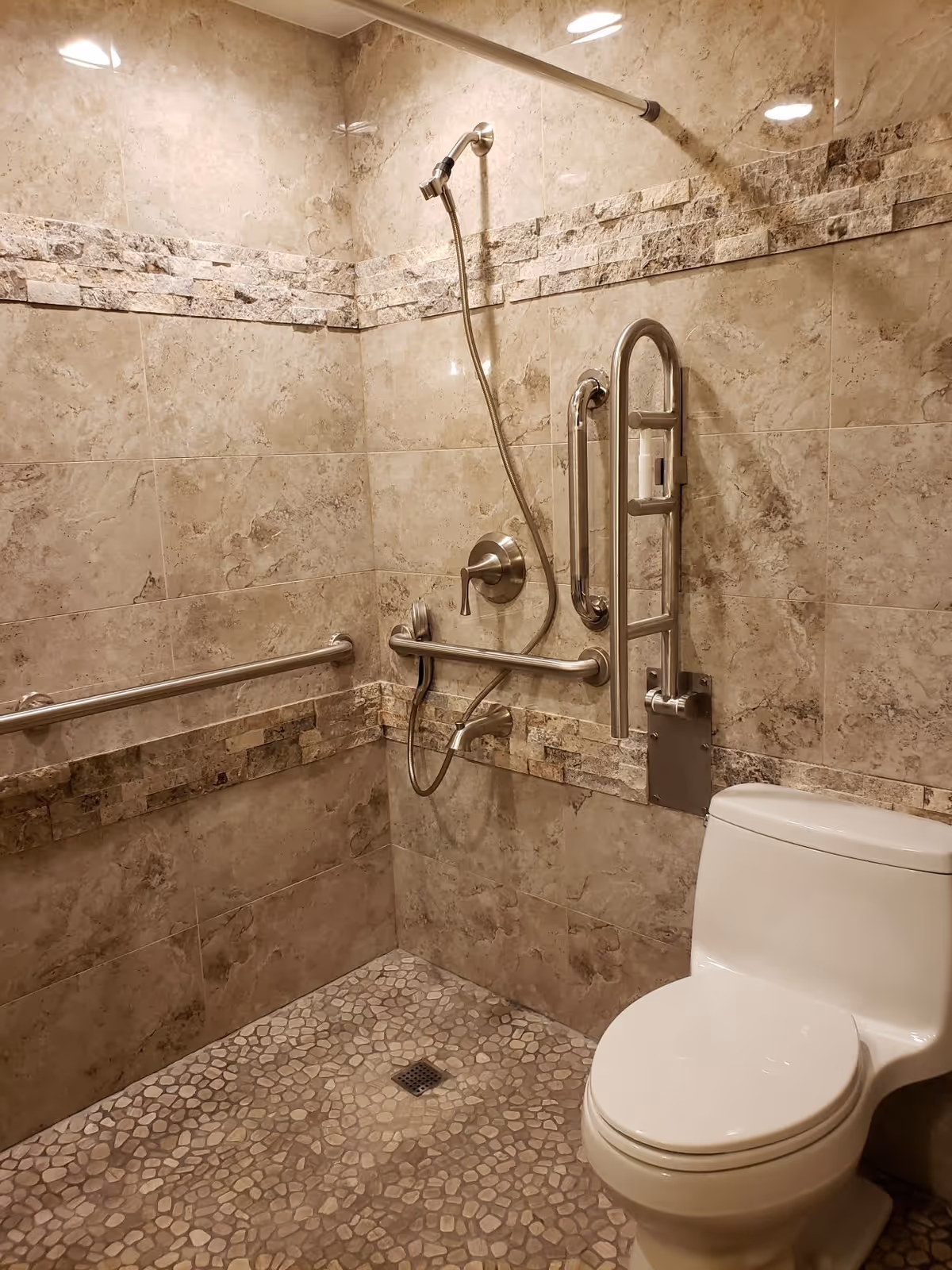 A bathroom with beige stone tile walls and pebble-textured floor. It features a white toilet and a shower area with a handheld showerhead, multiple stainless steel grab bars for accessibility, and a wall-mounted soap dispenser.