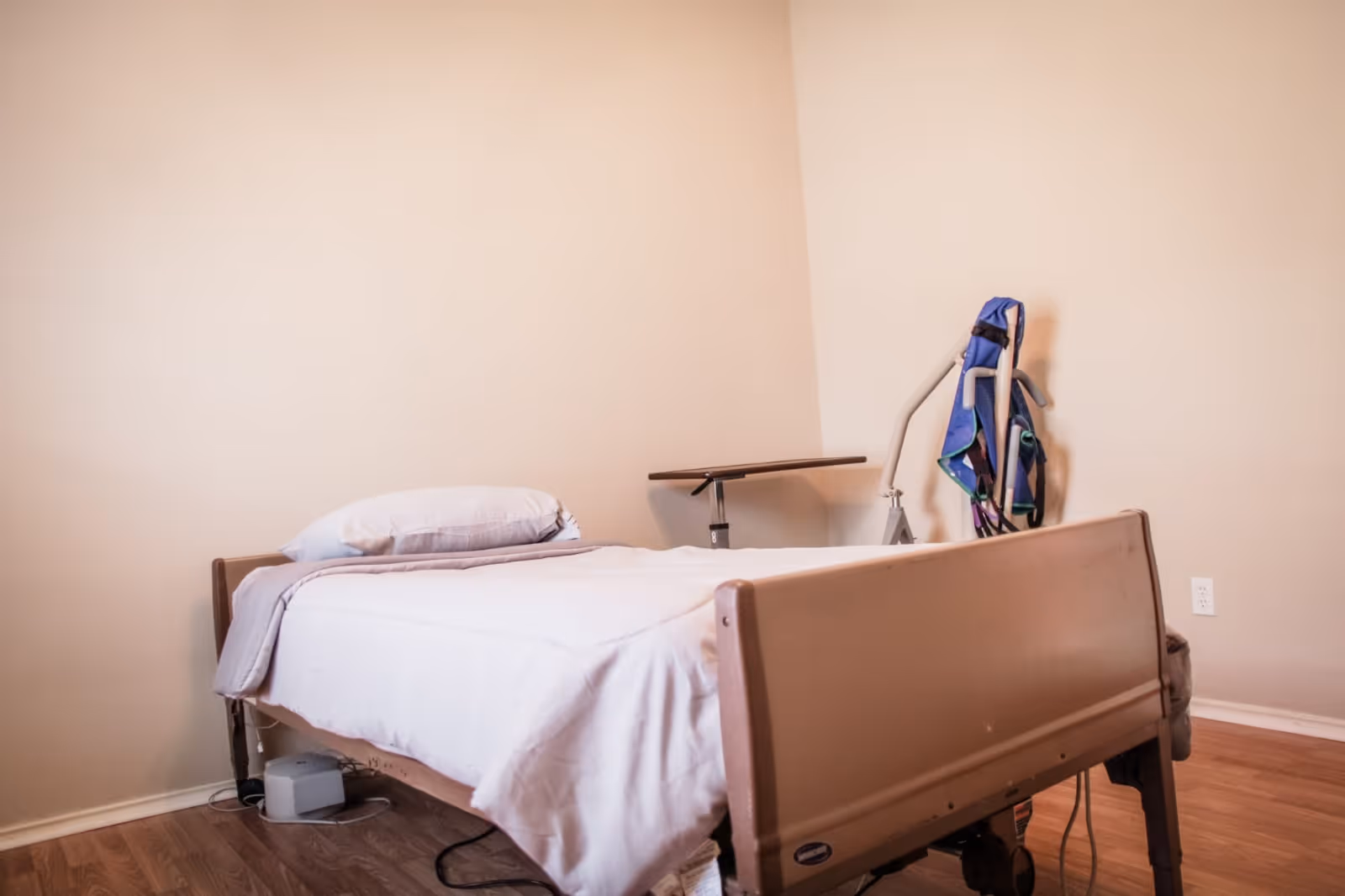 A simple patient bedroom with a hospital-style bed, overbed table, and a sling lift against a beige wall.