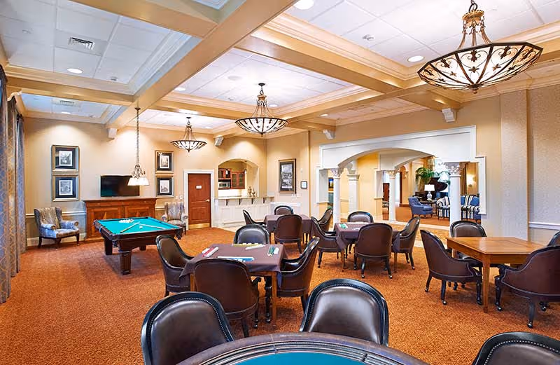 A spacious common area in a senior living facility featuring multiple tables with chairs arranged for games or socializing, a pool table, elegant chandeliers, and warm beige walls with framed artwork. The room has carpeted floors and an open archway leading to another seating area with sofas and plants.