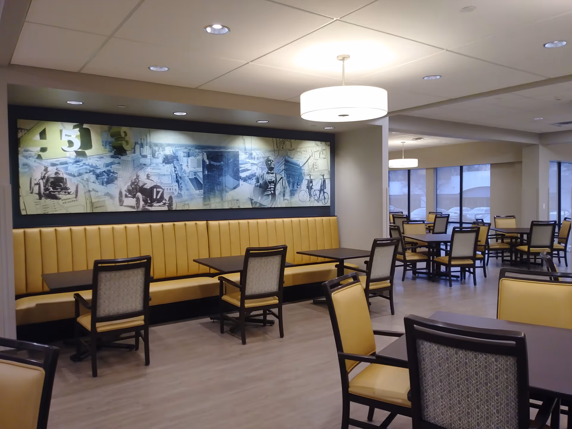 Interior view of a dining room with multiple tables and chairs. The chairs have yellow cushions and dark wooden frames. There is a long yellow cushioned bench along one wall with a large mural above it depicting historical images and numbers. The room has large windows letting in natural light and modern ceiling lights.