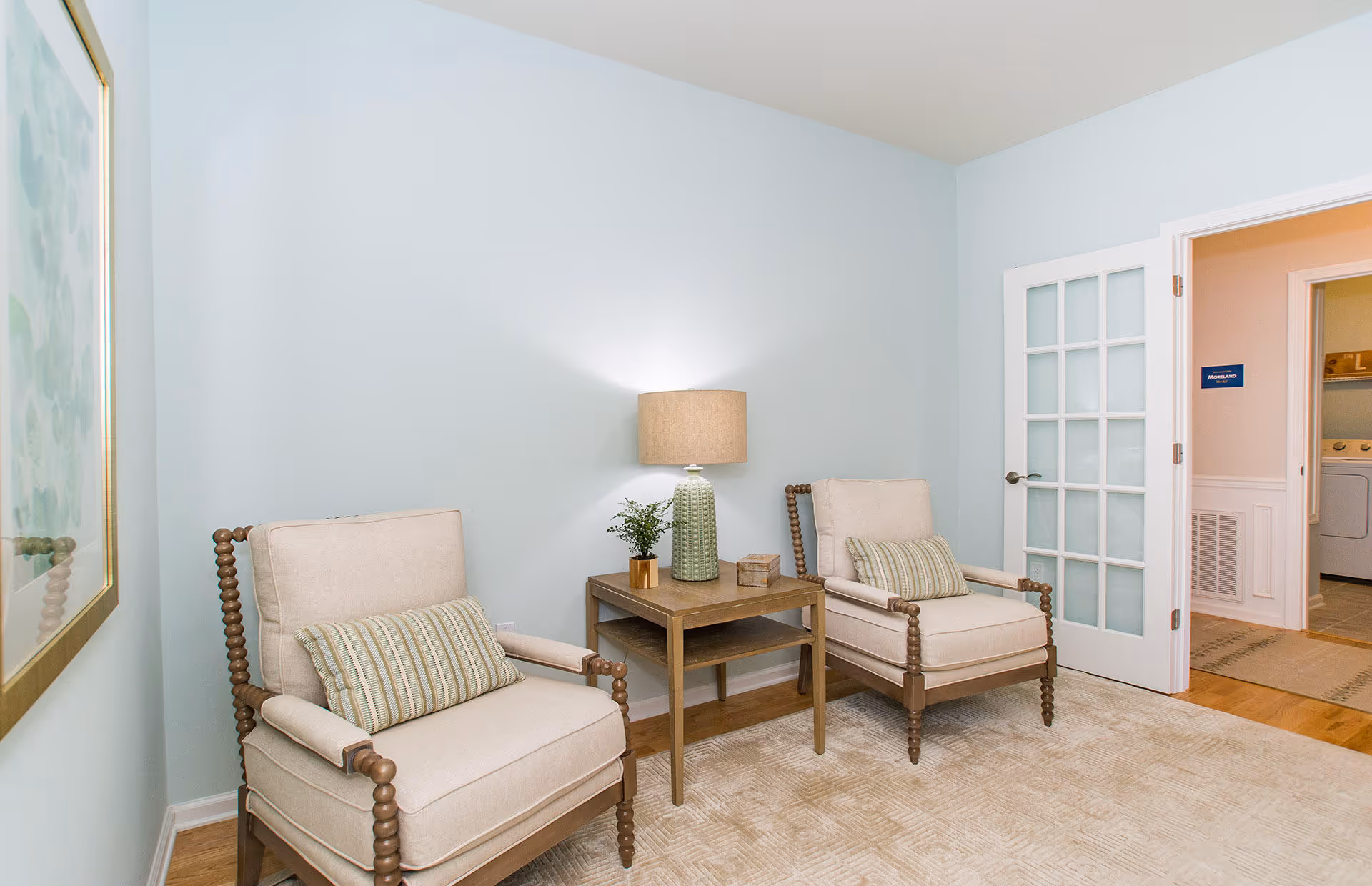 A cozy sitting area with two beige cushioned armchairs featuring wooden spindle arms and legs, separated by a wooden side table with a green ceramic lamp, a small plant, and a decorative box. The walls are painted light blue, and there is a framed artwork on the left wall. A glass-paneled door on the right leads to a laundry room with a washer and dryer visible.
