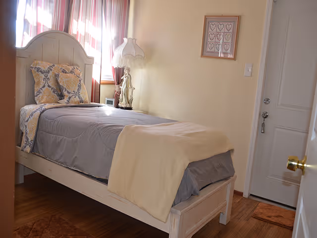 A small bedroom with a single bed featuring a white wooden frame, gray bedding, and decorative pillows. A vintage-style lamp with a figurine base is on a small table next to the bed. Sunlight filters through a window with striped curtains. A framed picture with heart designs hangs on the wall, and a white door with a silver doorknob is visible.