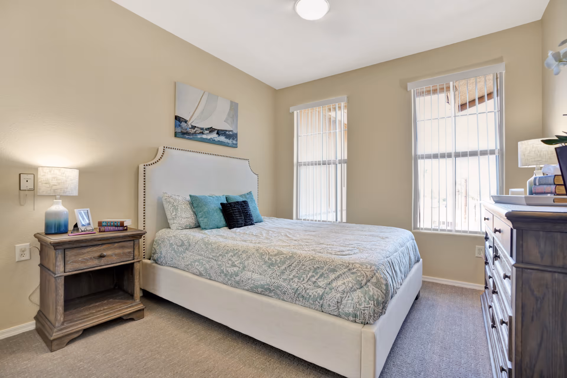 A neatly made bed with a light-colored upholstered headboard and patterned bedding in a bedroom. There are two windows with vertical blinds letting in natural light. On the left side of the bed is a wooden nightstand with a lamp, a framed photo, and some books. On the right side of the room is a wooden dresser with a lamp and decorative items on top. A painting of a sailboat hangs on the wall above the bed.