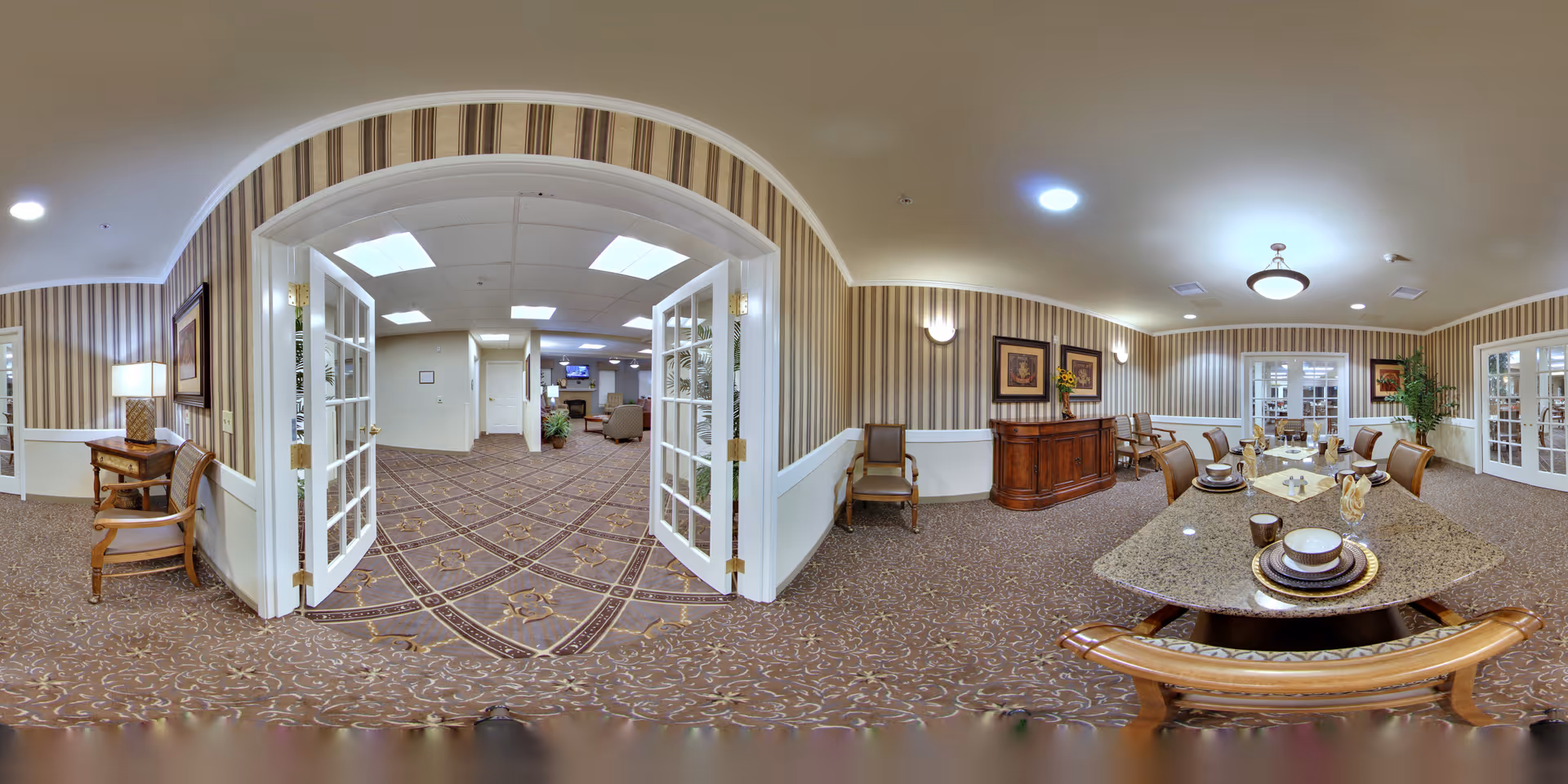 A spacious, well-lit interior room in a senior living facility featuring a large dining table set with plates and cups on the right side, several chairs, and a wooden sideboard against the wall. The room has patterned carpet and striped wallpaper with framed artwork. Double glass doors open into an adjacent lounge area with more seating and a television.