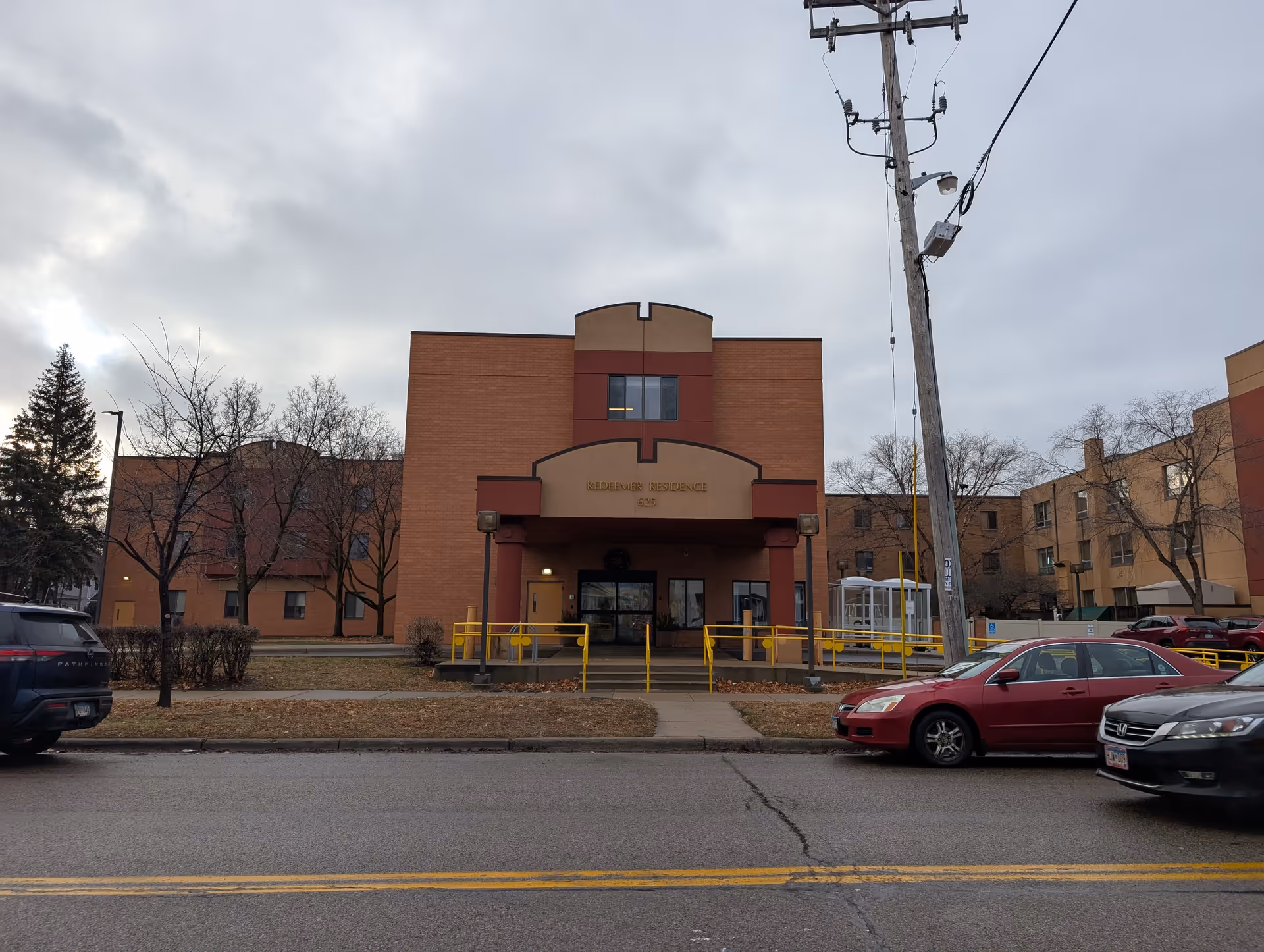 Front exterior of a brick building labeled Redeemer Residence with a covered entrance, yellow railings, and cars parked along the street.