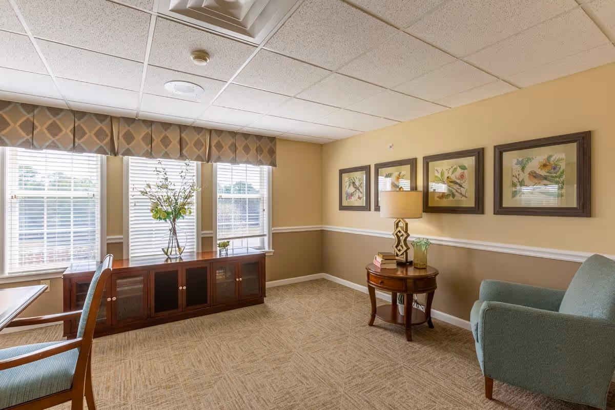 A cozy sitting area in a senior living facility with beige walls and carpeted floor. The room features three large windows with blinds and a patterned valance, a wooden cabinet with glass doors beneath the windows, a small round wooden side table with a lamp, books, and a glass vase, and a comfortable upholstered armchair. Three framed bird-themed artworks hang on the wall above the side table.