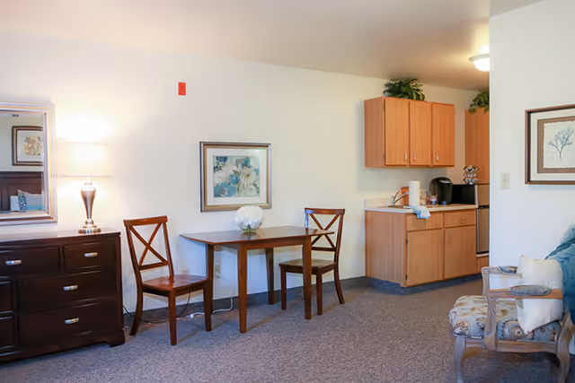 A cozy senior living area featuring a small wooden dining table with two chairs, a wooden dresser with a lamp and mirror, a kitchenette with wooden cabinets, and a cushioned armchair with a pillow. The walls are decorated with framed floral artwork.