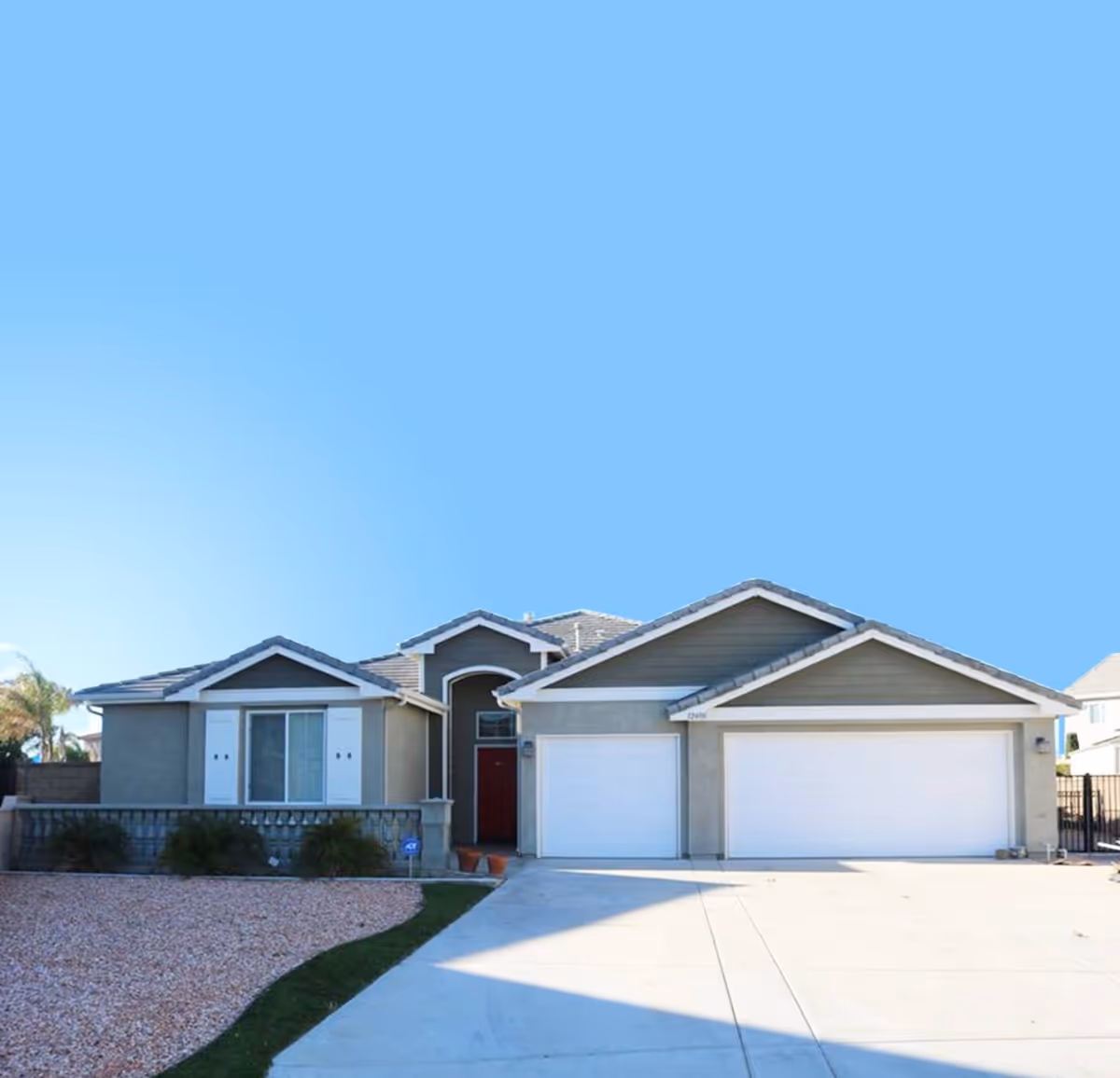 Front exterior of a single-story suburban house with a three-car garage, driveway, and gravel front yard under a clear blue sky.