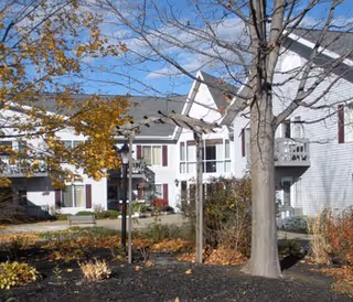 Exterior view of a senior living facility with white siding buildings, multiple windows, and balconies. There is a tree with autumn leaves and landscaped garden beds in the foreground under a blue sky with scattered clouds.
