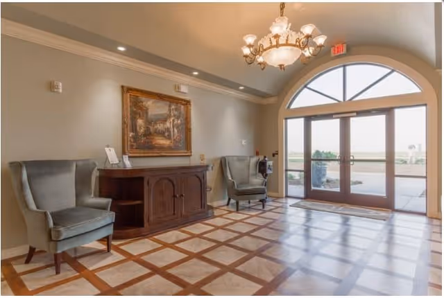 A spacious and well-lit entrance lobby with a large arched glass door letting in natural light. The floor features a checkered pattern of tiles and wood. Two gray upholstered armchairs flank a wooden console table against the wall, which is decorated with a framed painting. A chandelier hangs from the ceiling, and there is an exit sign above the door.