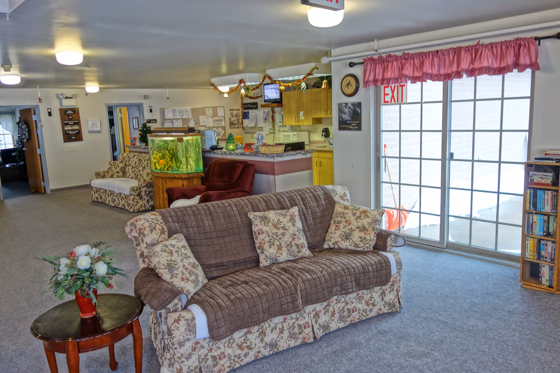 A cozy living room area in a senior living facility with a floral patterned sofa covered by a brown quilted throw and matching pillows. There is a small round wooden table with a vase of flowers in front of the sofa. In the background, there is a fish tank on a wooden stand, a red armchair, and a kitchenette with wooden cabinets. A sliding glass door with pink curtains leads to an outdoor area. A bookshelf filled with books is visible on the right side.