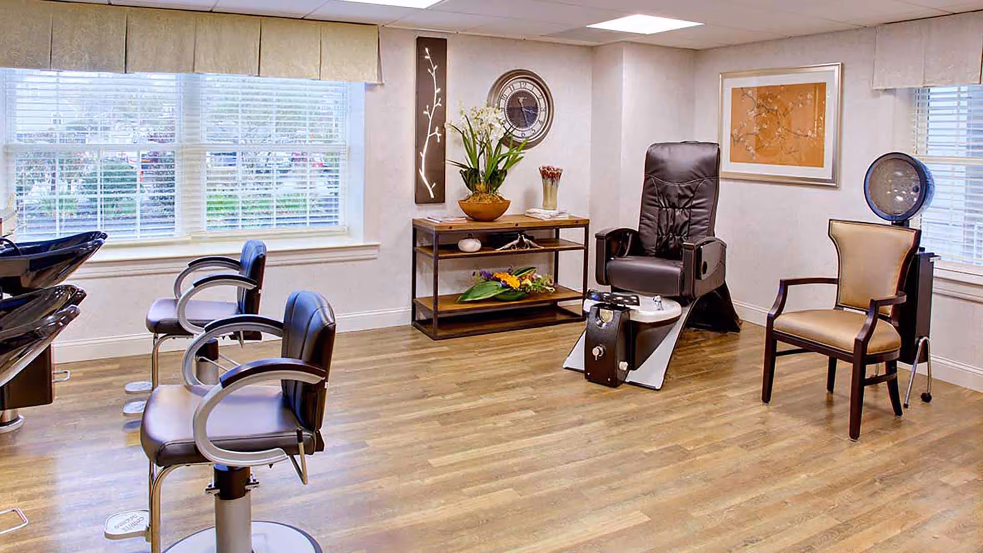 Interior of a salon area with two black salon chairs in front of hair washing stations, a black pedicure chair with foot spa, a beige armchair, a wooden shelf with decorative plants and flowers, a wall clock, and framed artwork on the walls. The room has large windows with blinds and light-colored walls and flooring.