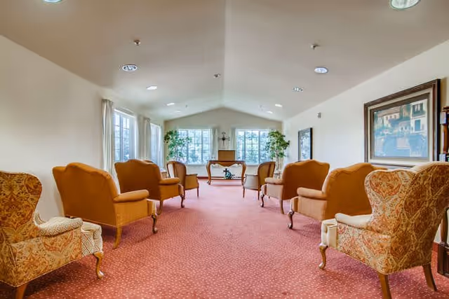 A spacious, well-lit common area with multiple upholstered armchairs arranged in two rows facing each other. The room has large windows at the far end letting in natural light, potted plants near the windows, and framed artwork on the walls. The floor is covered with a red patterned carpet.