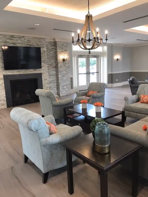 A bright and spacious living room area with light-colored armchairs arranged around dark wooden coffee tables. The room features a stone fireplace with a mounted flat-screen TV above it, a chandelier hanging from the ceiling, and double glass doors letting in natural light. The decor includes small orange flower arrangements and a decorative glass jar on the table in the foreground.