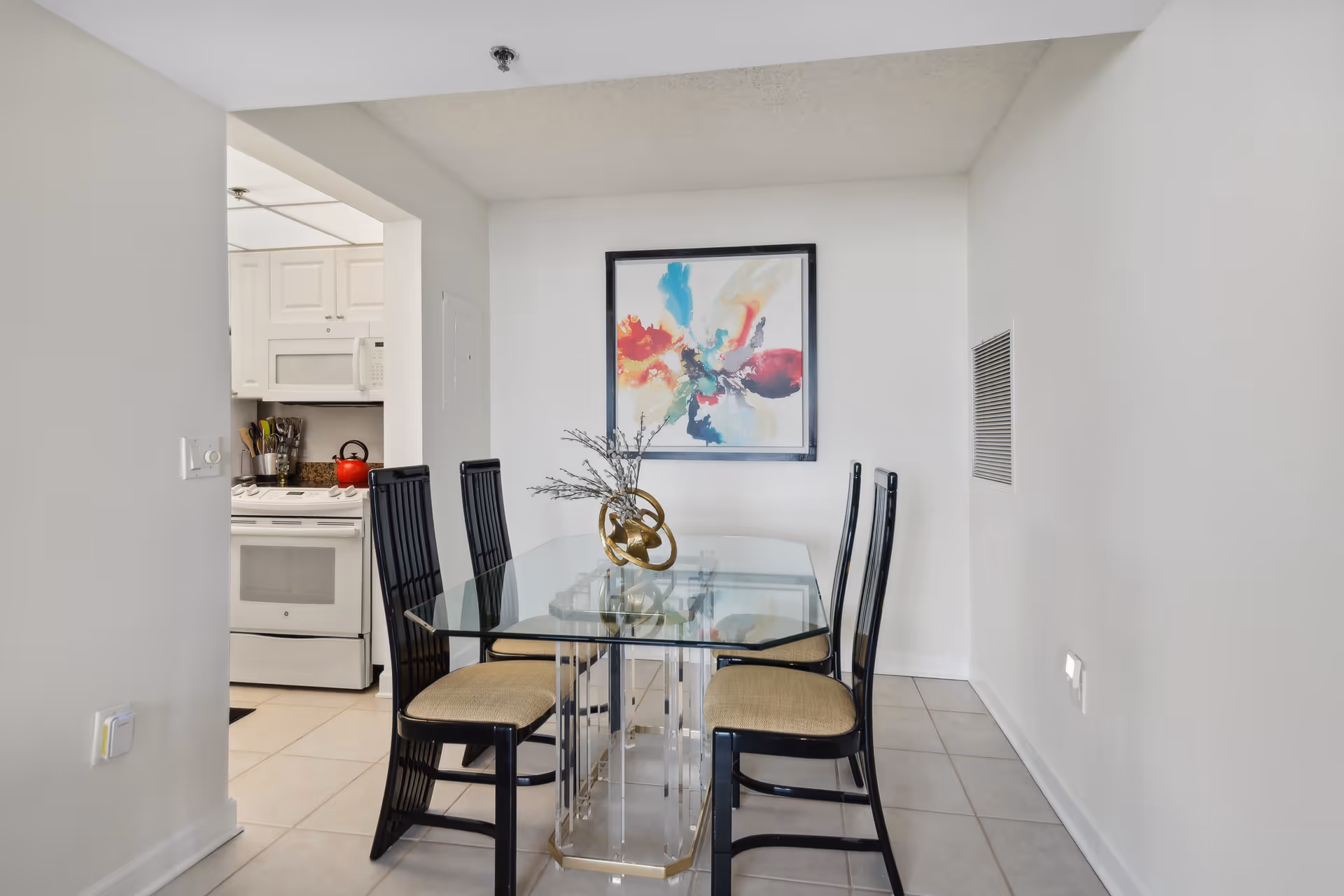 A dining area with a glass-top table and four black chairs with beige cushions. A decorative centerpiece with branches is on the table. On the wall behind the table is a colorful abstract painting. To the left, there is a partial view of a kitchen with white cabinets, a white stove, and a red kettle on the stove.