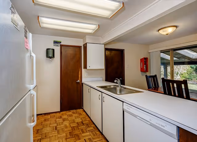 Interior view of a kitchen with white cabinets and countertops, a double sink, a dishwasher, a refrigerator, and wooden doors. There is a dining area with wooden chairs visible through a sliding glass door leading to an outdoor space.