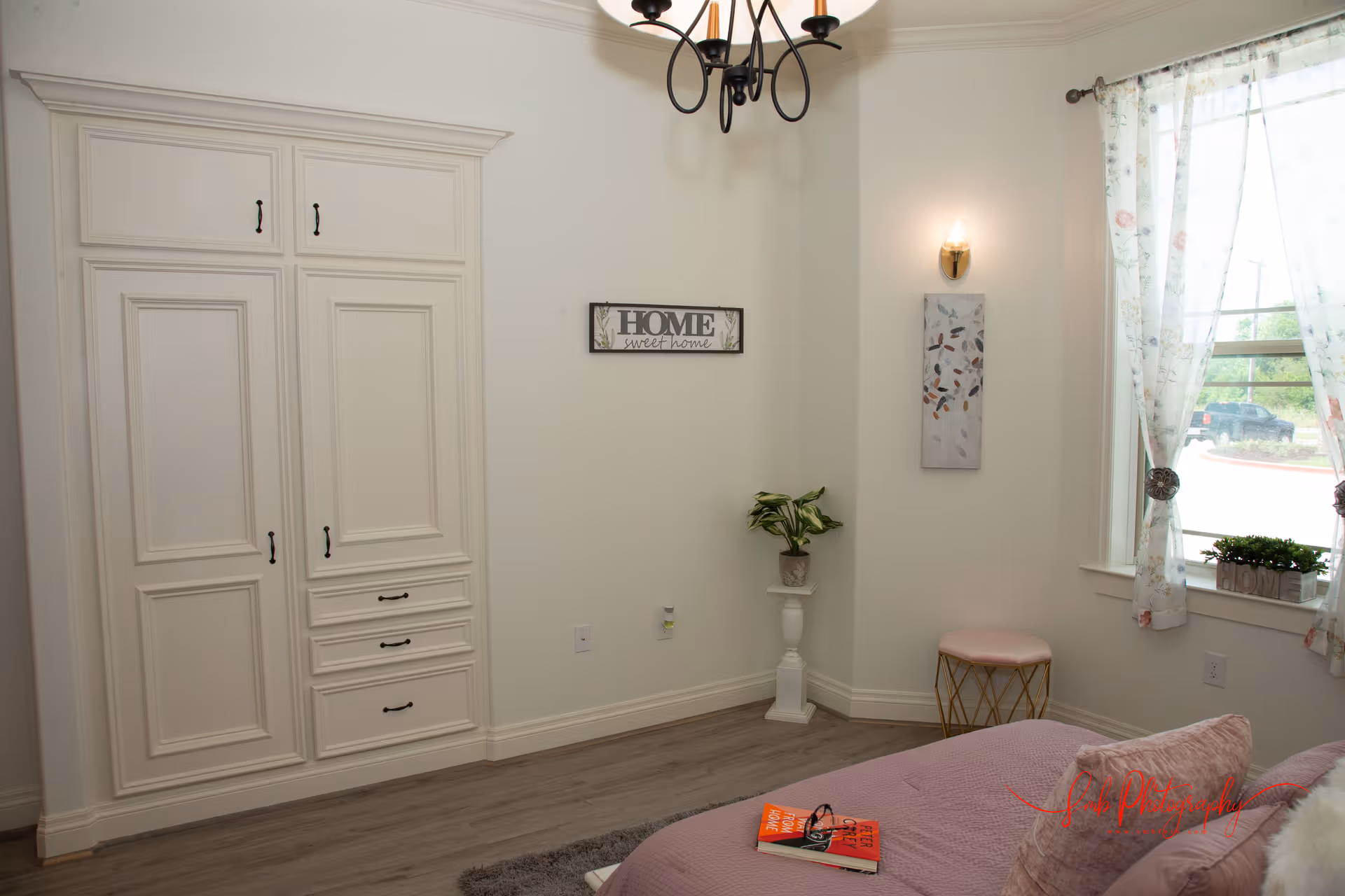 A cozy bedroom with a white built-in wardrobe, a bed with pink bedding and pillows, a small round stool, a potted plant on a white pedestal, wall art, a chandelier, and a window with floral curtains letting in natural light.