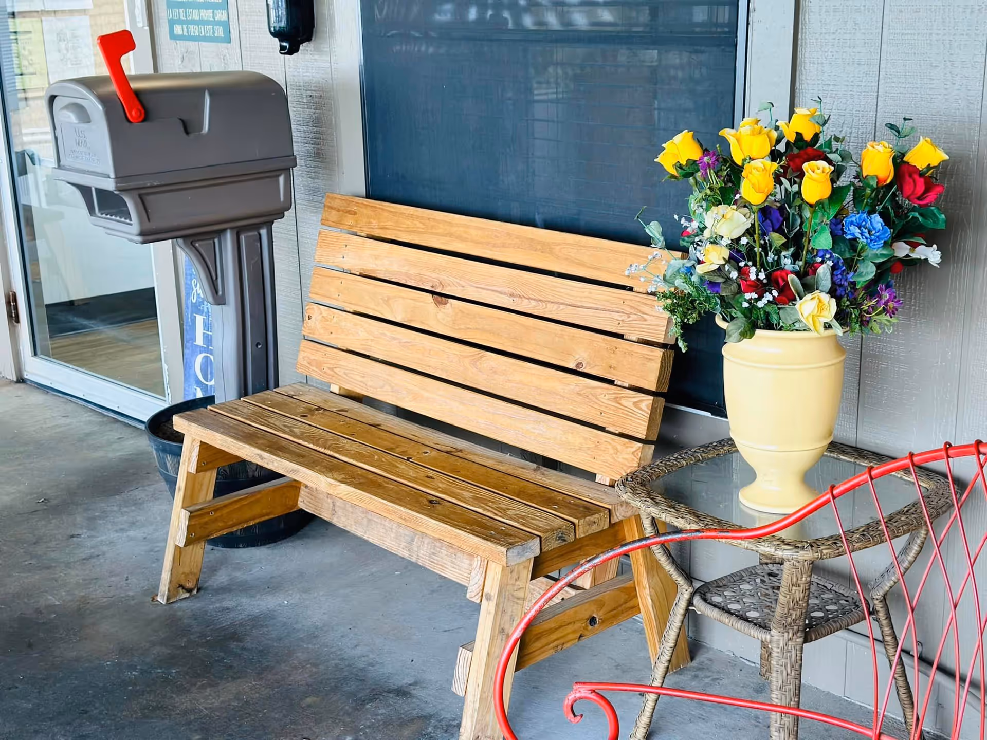 Wooden bench and small table holding a vase of colorful flowers next to a mailbox on a building porch.