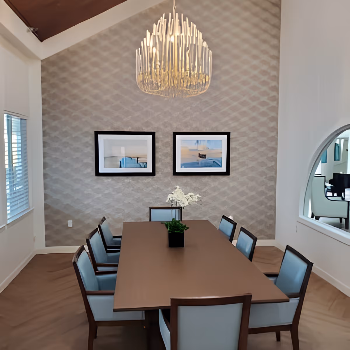 A modern dining room with a long wooden table surrounded by chairs, a chandelier overhead, framed artwork on a patterned accent wall, and a small plant centerpiece.