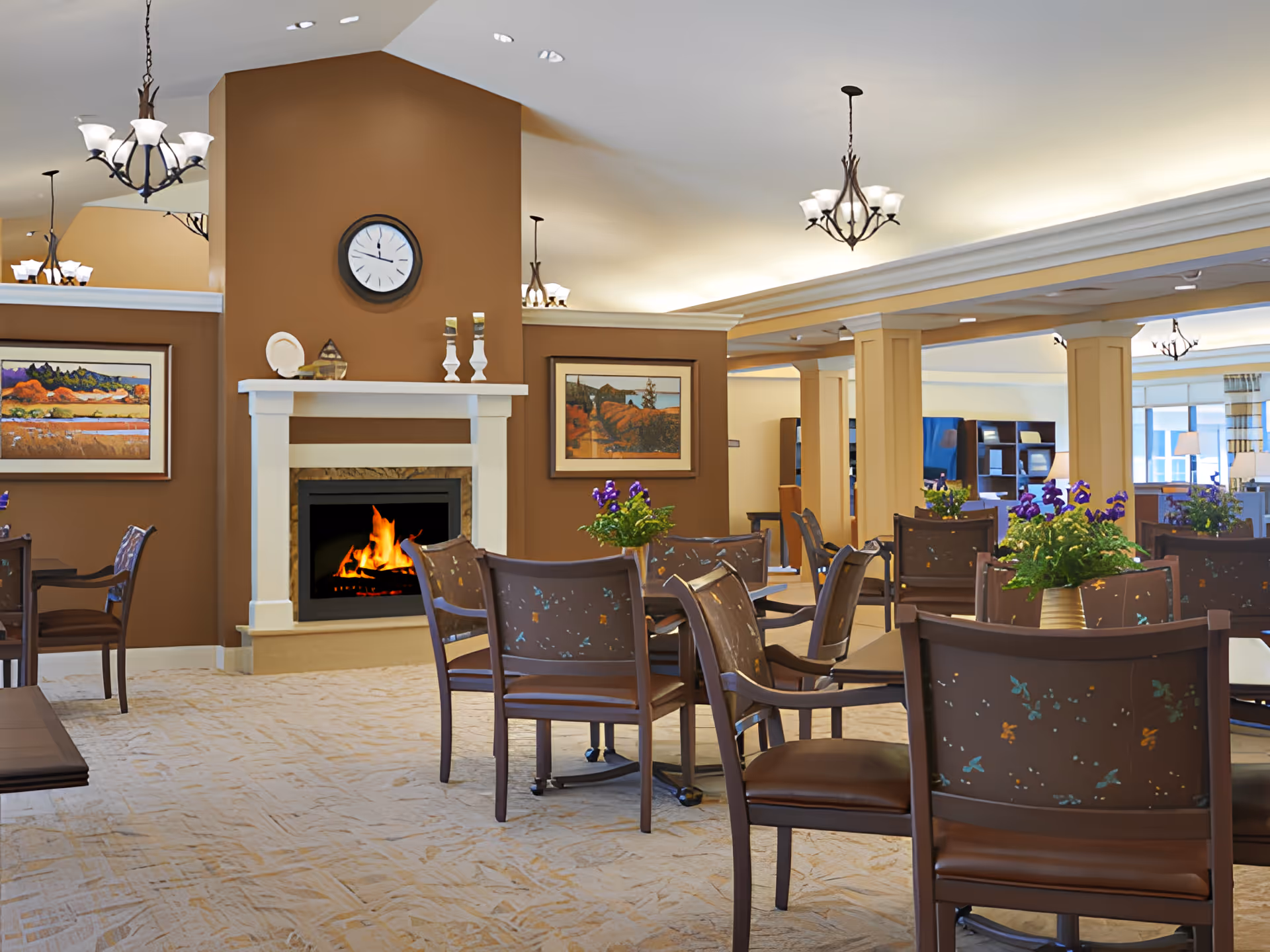 Communal dining/lounge room with multiple tables and chairs, a lit fireplace, wall art, and hanging chandeliers.