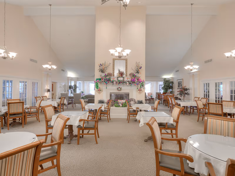 A spacious dining room with multiple tables covered with white tablecloths and surrounded by wooden chairs with striped cushions. The room features a central fireplace decorated with flowers and garlands, high vaulted ceilings with hanging chandeliers, and large windows allowing natural light to fill the space.