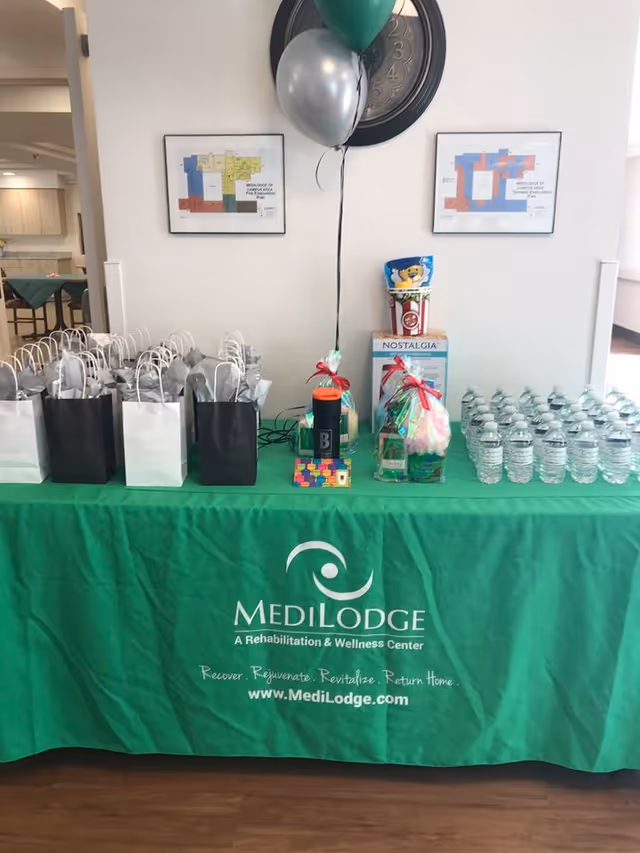 A table covered with a green tablecloth displaying the Medilodge logo and website. On the table are multiple black and white gift bags, two balloons (one green and one silver), several bottles of water, and some gift items including a popcorn container and a Nostalgia brand item. Behind the table on the wall are two framed floor plans and a large clock.