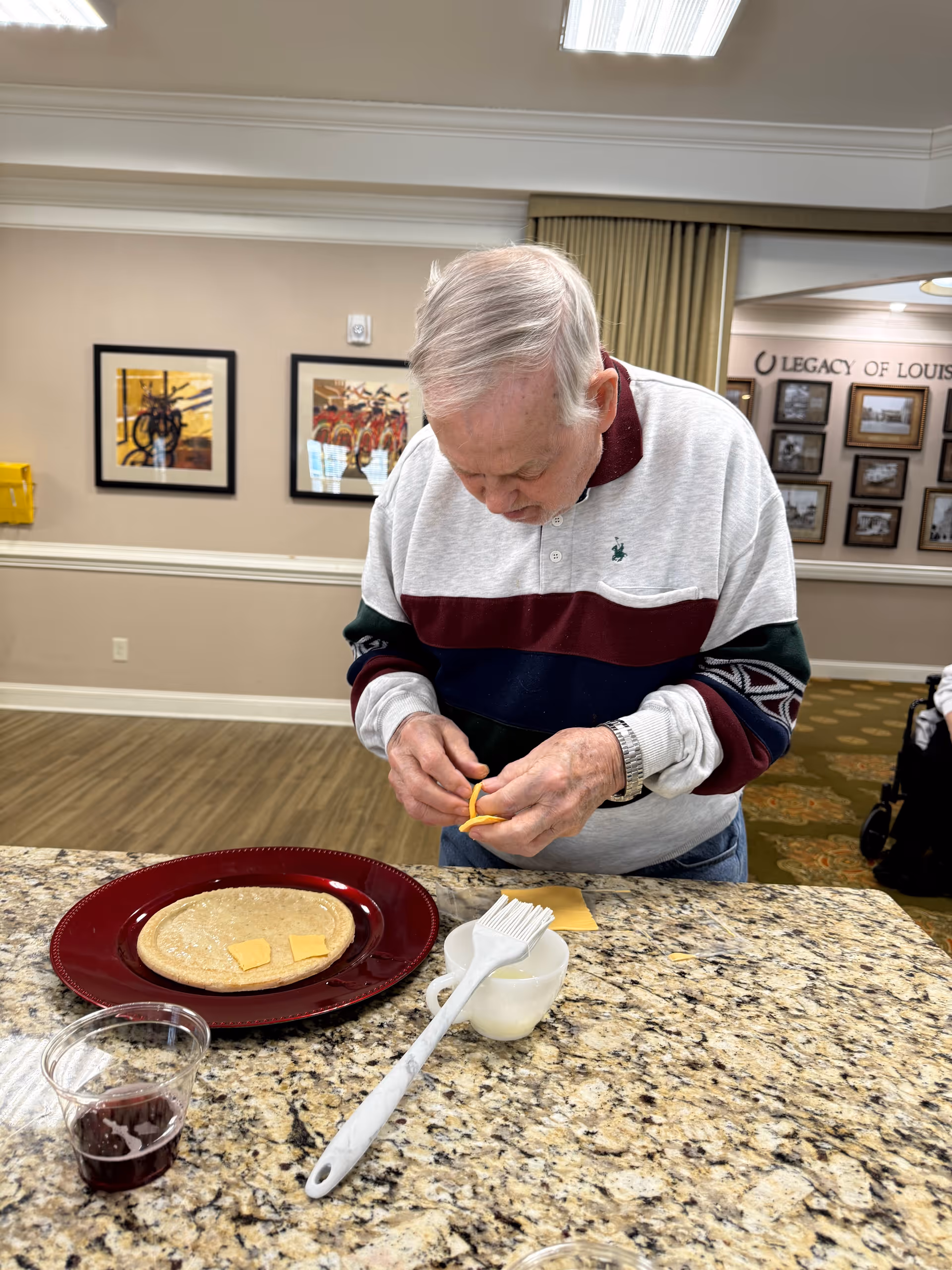An elderly man standing at a granite countertop in a communal indoor space, preparing food by placing small slices of cheese on a tortilla on a red plate. There is a small cup with a white brush and a clear cup with a dark liquid on the counter. The background shows framed pictures on beige walls and a sign that reads 'Legacy of Louis'.