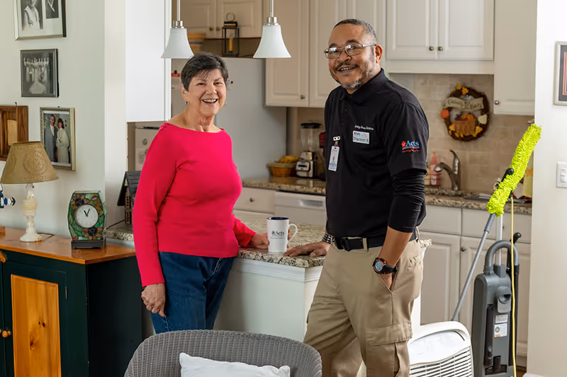 An elderly woman in a red sweater and blue jeans stands smiling next to a man in a black polo shirt and khaki pants in a kitchen area. The man is wearing glasses and a name badge. The kitchen has white cabinets, a granite countertop, and various appliances. A vacuum cleaner and cleaning tools are visible in the background.