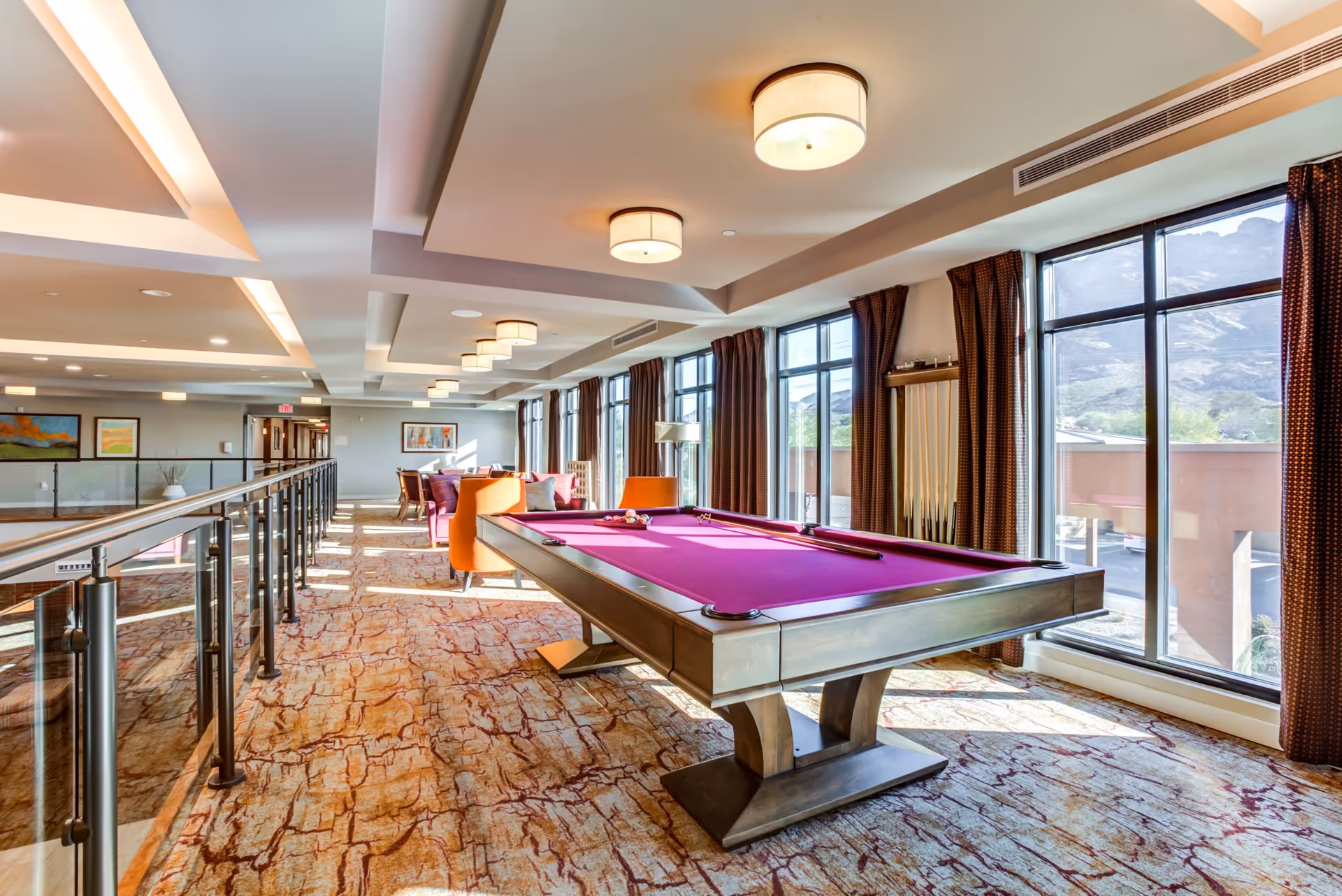 A bright and spacious recreational room with a modern pool table featuring a purple felt surface. The room has large floor-to-ceiling windows with brown curtains, allowing natural light to fill the space and offering a view of mountains outside. There are several ceiling lights, a patterned carpet, and comfortable seating areas with orange and purple chairs in the background.