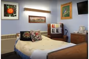 A single bed with floral pillows and a beige blanket in a room with light blue walls. There is a wooden nightstand with a lamp, a TV mounted on the wall, and several framed pictures hanging on the walls. A cushioned chair with a decorative throw is placed next to the bed.