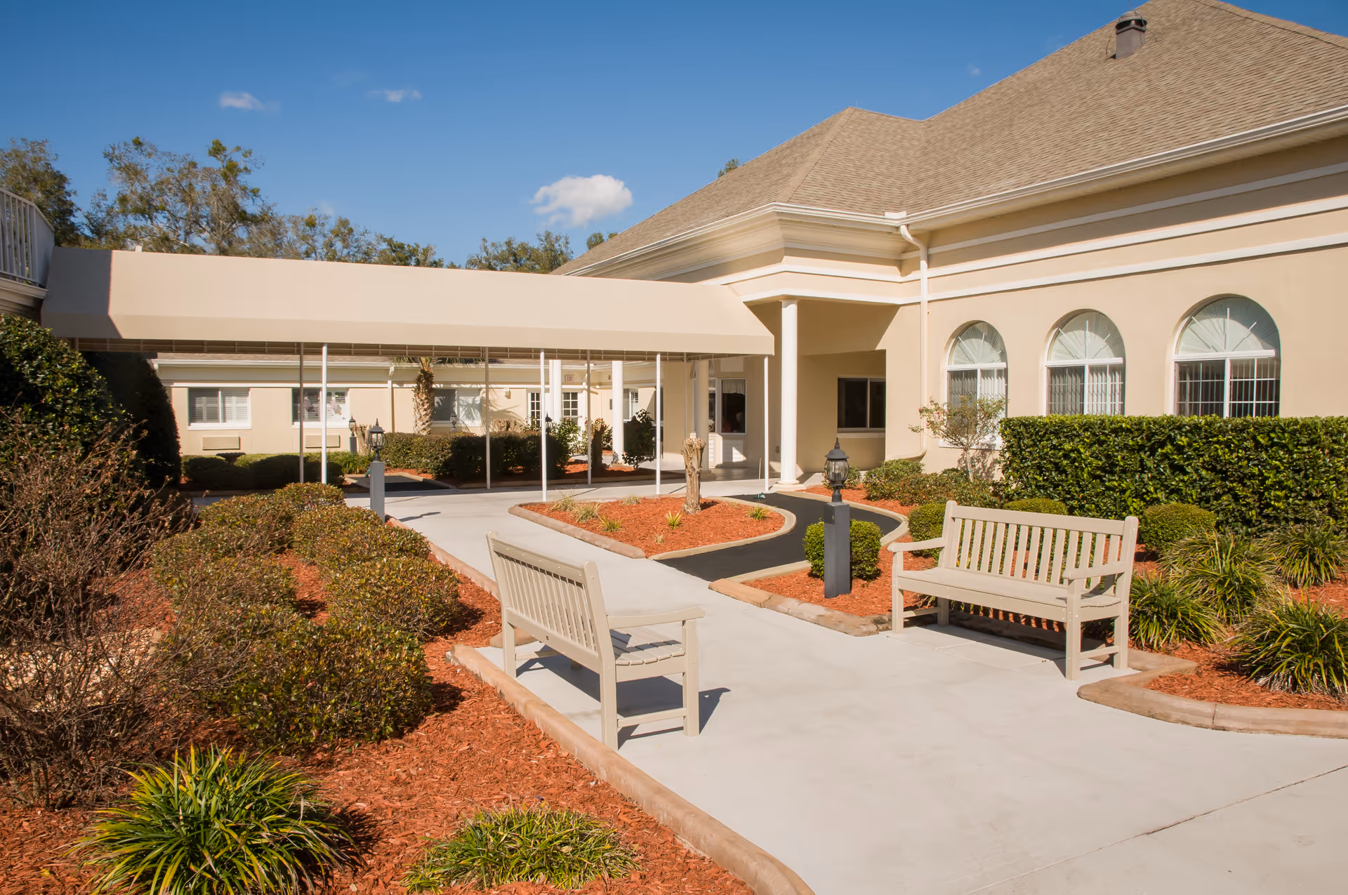 Outdoor view of the entrance area of a senior living facility with a covered walkway, beige building with arched windows, landscaped garden beds with bushes and mulch, and two wooden benches on a concrete path under a clear blue sky.