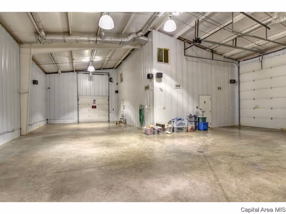 Spacious empty metal-walled garage or warehouse interior with concrete floor, overhead lights, roll-up doors and storage bins against a wall.