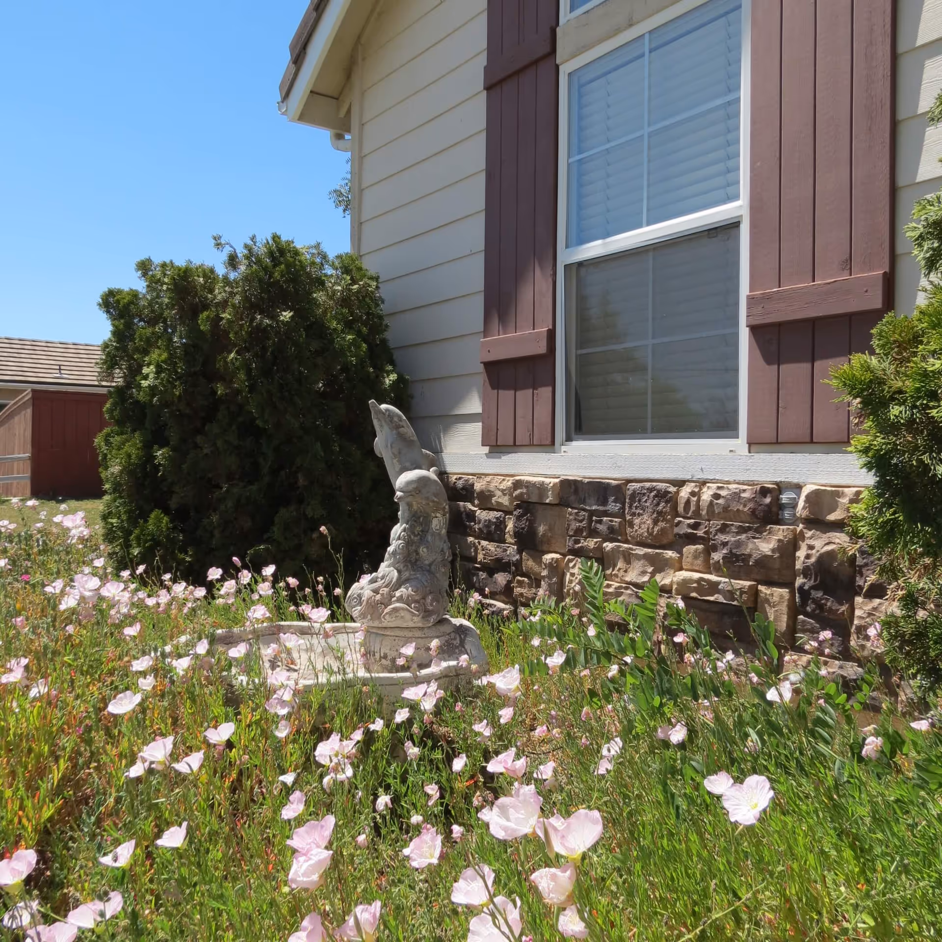 Outdoor garden area next to a building with beige siding and stone accents. There are pink flowers in bloom, green bushes, and a decorative stone bird sculpture in the garden bed under a window with brown shutters.