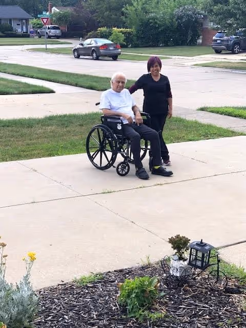 An elderly man sitting in a wheelchair on a sidewalk outside, accompanied by a woman standing next to him. They are in a residential neighborhood with houses, cars, and greenery visible in the background.