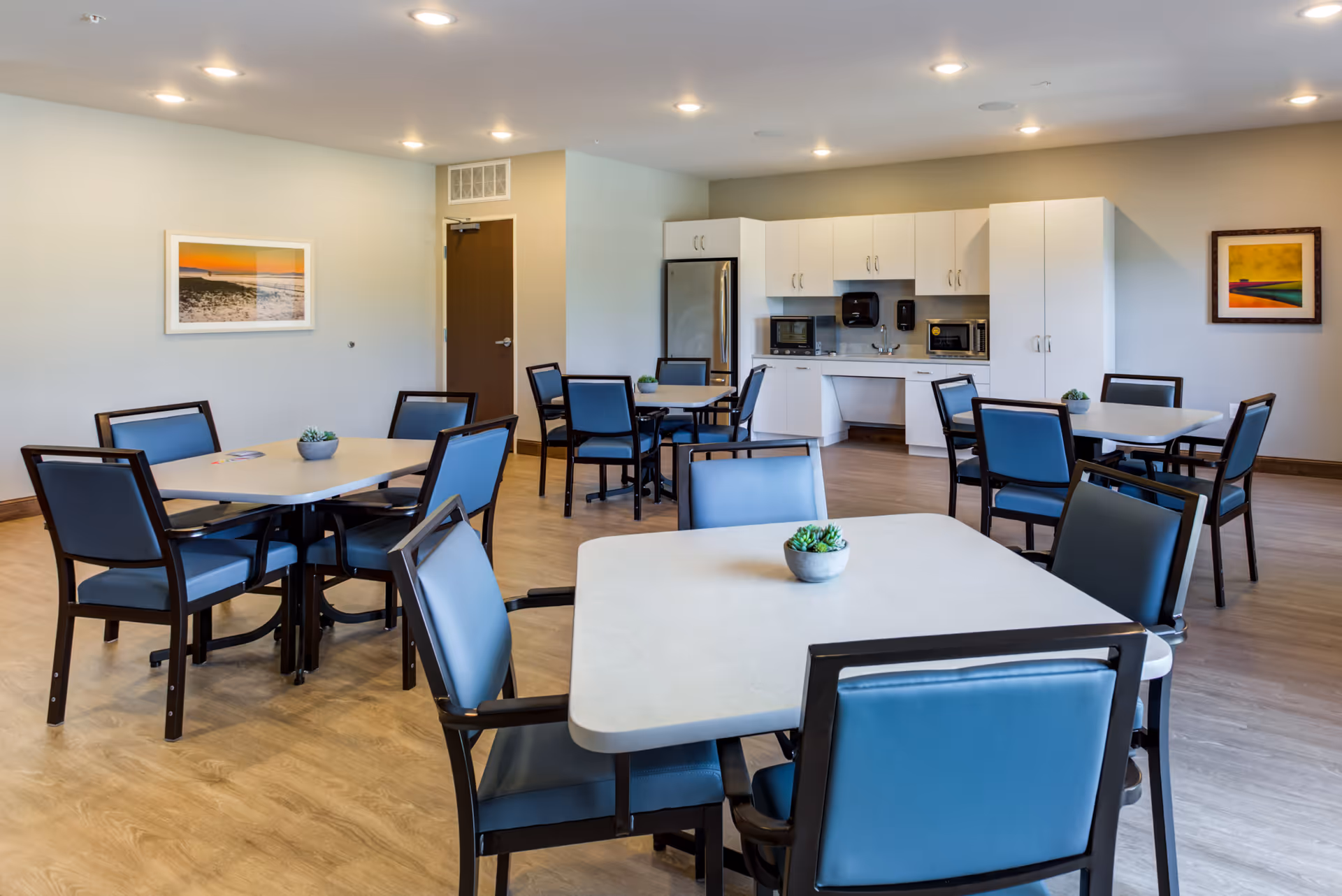 A bright dining area in a senior living facility featuring several square tables with blue cushioned chairs. Each table has a small potted plant centerpiece. The room has light wood flooring, white walls with framed artwork, and a kitchenette area with white cabinets, a refrigerator, microwave ovens, and a sink.