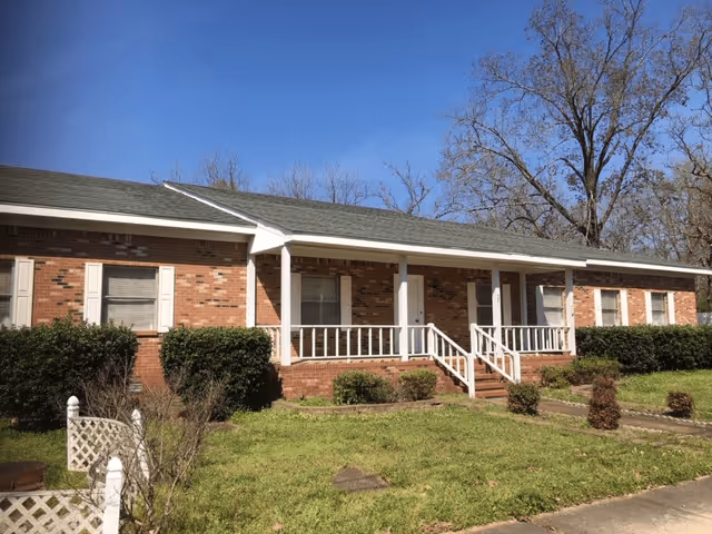 Single-story brick building with a green roof and white porch railings, surrounded by green grass and bushes under a clear blue sky.