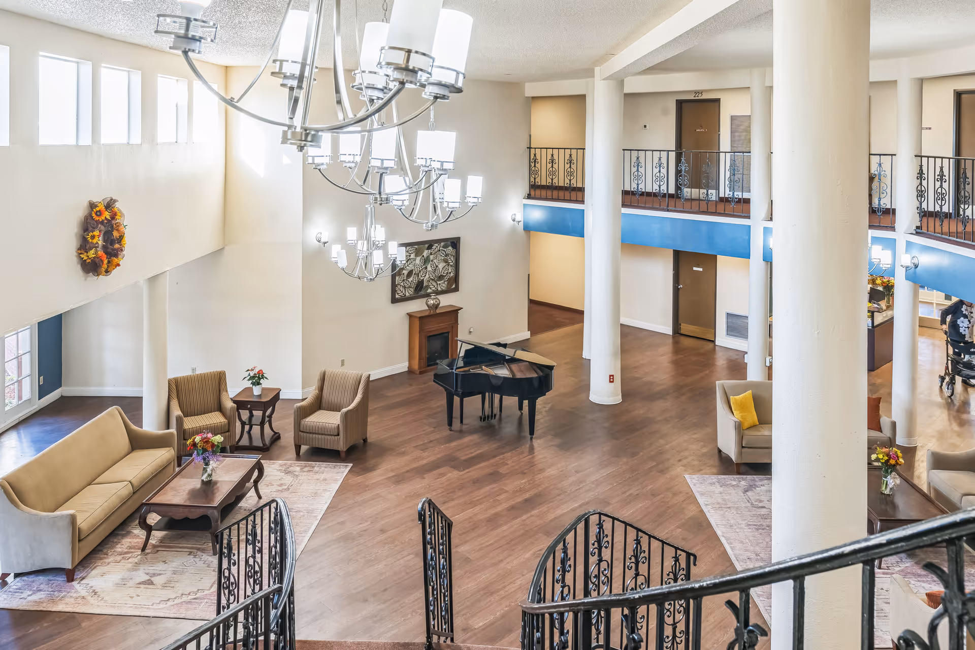 Spacious senior living facility common area with high ceilings, large windows, and modern chandeliers. The room features a grand piano, several seating areas with sofas and armchairs arranged around coffee tables, decorative rugs, and a fireplace. There is a second-floor balcony with ornate railings overlooking the area.